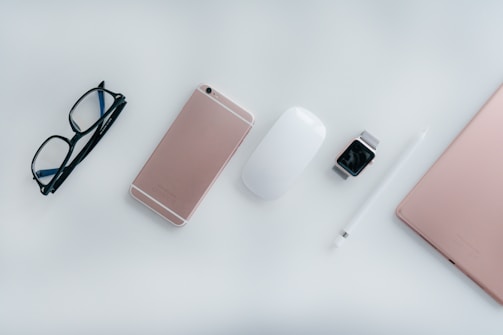 On a white surface, there's a neatly arranged collection of tech items and accessories. A pair of black glasses is positioned on the left. Next to the glasses lies a rose gold smartphone. To the right of the phone is a white computer mouse followed by a smart watch with a metallic band. Beside the watch, a white stylus pen is aligned parallel to a rose gold laptop's side edge.