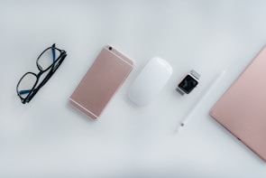 On a white surface, there's a neatly arranged collection of tech items and accessories. A pair of black glasses is positioned on the left. Next to the glasses lies a rose gold smartphone. To the right of the phone is a white computer mouse followed by a smart watch with a metallic band. Beside the watch, a white stylus pen is aligned parallel to a rose gold laptop's side edge.