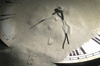 A clock partially buried in fine sand with Roman numerals partially visible. The clock hands are still prominent, lying on top of the sand, creating a curious blend of time and nature.