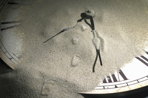 A clock partially buried in fine sand with Roman numerals partially visible. The clock hands are still prominent, lying on top of the sand, creating a curious blend of time and nature.