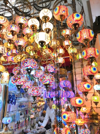 An indoor market stall features an array of vibrant, colorful mosaic lamps hanging closely together, each with intricate geometric patterns. A person is visible inside the stall, surrounded by various glass and metal items. The scene is rich in textures and colors, with warm illumination creating a vivid and inviting atmosphere.