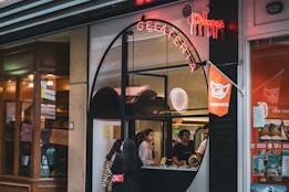A gelato shop with a distinctive arched window serves customers while displaying a neon sign above. Inside, two people stand at the counter, ready to assist. The exterior has a combination of modern and classic architectural elements. To the right, bright orange decor and signage are visible.