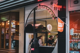 A gelato shop with a distinctive arched window serves customers while displaying a neon sign above. Inside, two people stand at the counter, ready to assist. The exterior has a combination of modern and classic architectural elements. To the right, bright orange decor and signage are visible.