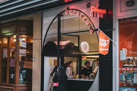 A gelato shop with a distinctive arched window serves customers while displaying a neon sign above. Inside, two people stand at the counter, ready to assist. The exterior has a combination of modern and classic architectural elements. To the right, bright orange decor and signage are visible.