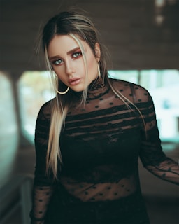 A woman with long, straight hair and hoop earrings is wearing a black, sheer, polka-dotted top. The background appears blurred, adding focus to her striking facial features and expression.