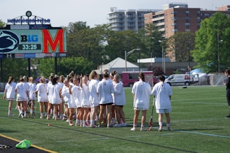 A group of female athletes in white sports attire are gathered on a grassy field, holding lacrosse sticks. In the background, there is a scoreboard displaying team logos and a timer. The setting is outdoors with buildings and trees visible beyond the field.