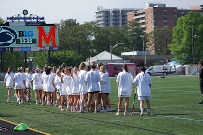 A group of female athletes in white sports attire are gathered on a grassy field, holding lacrosse sticks. In the background, there is a scoreboard displaying team logos and a timer. The setting is outdoors with buildings and trees visible beyond the field.