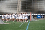 A group of women in white uniforms are running across a sports field, holding lacrosse sticks. They appear focused and determined. The background shows empty stadium bleachers and a blue 'Big' logo on a banner. There are lacrosse goals at either end of the field.