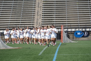 A group of women in white uniforms are running across a sports field, holding lacrosse sticks. They appear focused and determined. The background shows empty stadium bleachers and a blue 'Big' logo on a banner. There are lacrosse goals at either end of the field.
