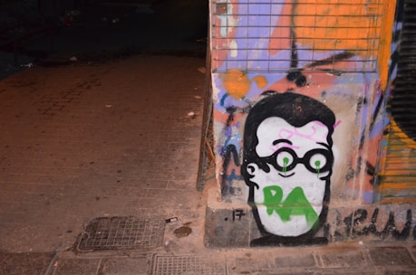 A graffiti artwork featuring a cartoonish face with black hair and glasses is painted on a wall. The eyes are emphasized with green circles, and the letters 'RA' are prominently displayed in green across the mouth area. The background includes abstract patterns in shades of orange, purple, and black. The scene is dimly lit, adding an urban feel to the street environment.