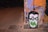 A graffiti artwork featuring a cartoonish face with black hair and glasses is painted on a wall. The eyes are emphasized with green circles, and the letters 'RA' are prominently displayed in green across the mouth area. The background includes abstract patterns in shades of orange, purple, and black. The scene is dimly lit, adding an urban feel to the street environment.