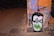 A graffiti artwork featuring a cartoonish face with black hair and glasses is painted on a wall. The eyes are emphasized with green circles, and the letters 'RA' are prominently displayed in green across the mouth area. The background includes abstract patterns in shades of orange, purple, and black. The scene is dimly lit, adding an urban feel to the street environment.