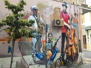 A vibrant mural on an urban wall features three musicians. One plays the saxophone, dressed in a white outfit and hat. Another musician in a red shirt stands with a large double bass. The third sits cross-legged, holding a string instrument. The mural is surrounded by an air conditioning unit, wires, and a bicycle parked below. A small tree grows nearby, adding greenery to the scene.
