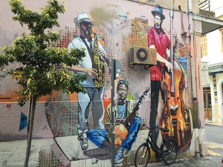 A vibrant mural on an urban wall features three musicians. One plays the saxophone, dressed in a white outfit and hat. Another musician in a red shirt stands with a large double bass. The third sits cross-legged, holding a string instrument. The mural is surrounded by an air conditioning unit, wires, and a bicycle parked below. A small tree grows nearby, adding greenery to the scene.