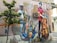 A vibrant mural on an urban wall features three musicians. One plays the saxophone, dressed in a white outfit and hat. Another musician in a red shirt stands with a large double bass. The third sits cross-legged, holding a string instrument. The mural is surrounded by an air conditioning unit, wires, and a bicycle parked below. A small tree grows nearby, adding greenery to the scene.