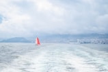A small sailboat with a vibrant red sail is navigating through the water, leaving ripples behind. In the distance, a larger cruise ship is docked near a coastline lined with buildings. The sky is overcast with thick clouds, creating a moody atmosphere over the water.