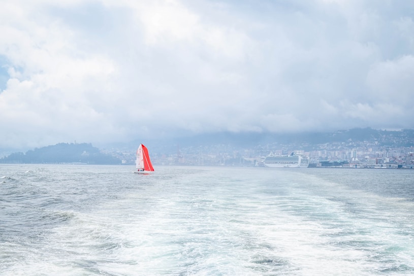 A small sailboat with a vibrant red sail is navigating through the water, leaving ripples behind. In the distance, a larger cruise ship is docked near a coastline lined with buildings. The sky is overcast with thick clouds, creating a moody atmosphere over the water.