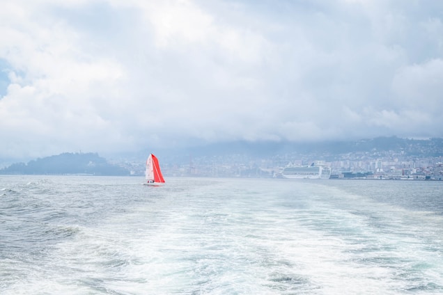 A small sailboat with a vibrant red sail is navigating through the water, leaving ripples behind. In the distance, a larger cruise ship is docked near a coastline lined with buildings. The sky is overcast with thick clouds, creating a moody atmosphere over the water.