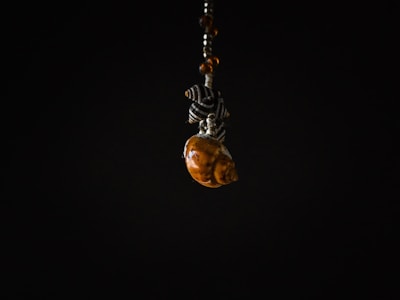 A small decorative object featuring a spiral seashell, intricately paired with black and white patterned beads. The object is suspended against a stark black background, creating a dramatic contrast that emphasizes the details of the seashell and beads.