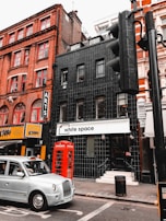 A street scene featuring a multi-story brick building with red window frames, a black-tiled building with a white sign that reads 'white space,' and a classic red telephone booth. A grey taxi is driving past, and there is a traffic light pole in the foreground.