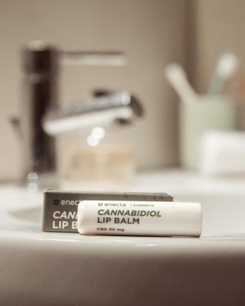 A tube of cannabidiol lip balm labeled CBD 50 mg is placed on a bathroom sink. The sink faucet is visible in the background along with blurred bathroom items such as a toothbrush holder.