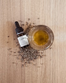A small glass bowl filled with yellow-green oil is placed on a wooden surface, surrounded by scattered hemp seeds. Next to the bowl is a dropper bottle labeled as CBD oil.