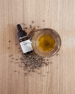 A small glass bowl filled with yellow-green oil is placed on a wooden surface, surrounded by scattered hemp seeds. Next to the bowl is a dropper bottle labeled as CBD oil.