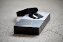 A black electronic device, possibly a fitness tracker or smartwatch, is resting on top of its packaging. The box is predominantly black and white with a large number three printed on it, suggesting a model or version number. The surface underneath is a beige carpet.