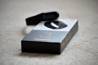 A black electronic device, possibly a fitness tracker or smartwatch, is resting on top of its packaging. The box is predominantly black and white with a large number three printed on it, suggesting a model or version number. The surface underneath is a beige carpet.