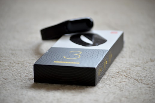 A black electronic device, possibly a fitness tracker or smartwatch, is resting on top of its packaging. The box is predominantly black and white with a large number three printed on it, suggesting a model or version number. The surface underneath is a beige carpet.