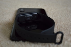 A smart band with a black strap lies resting on a smartphone, both placed on a textured beige surface.