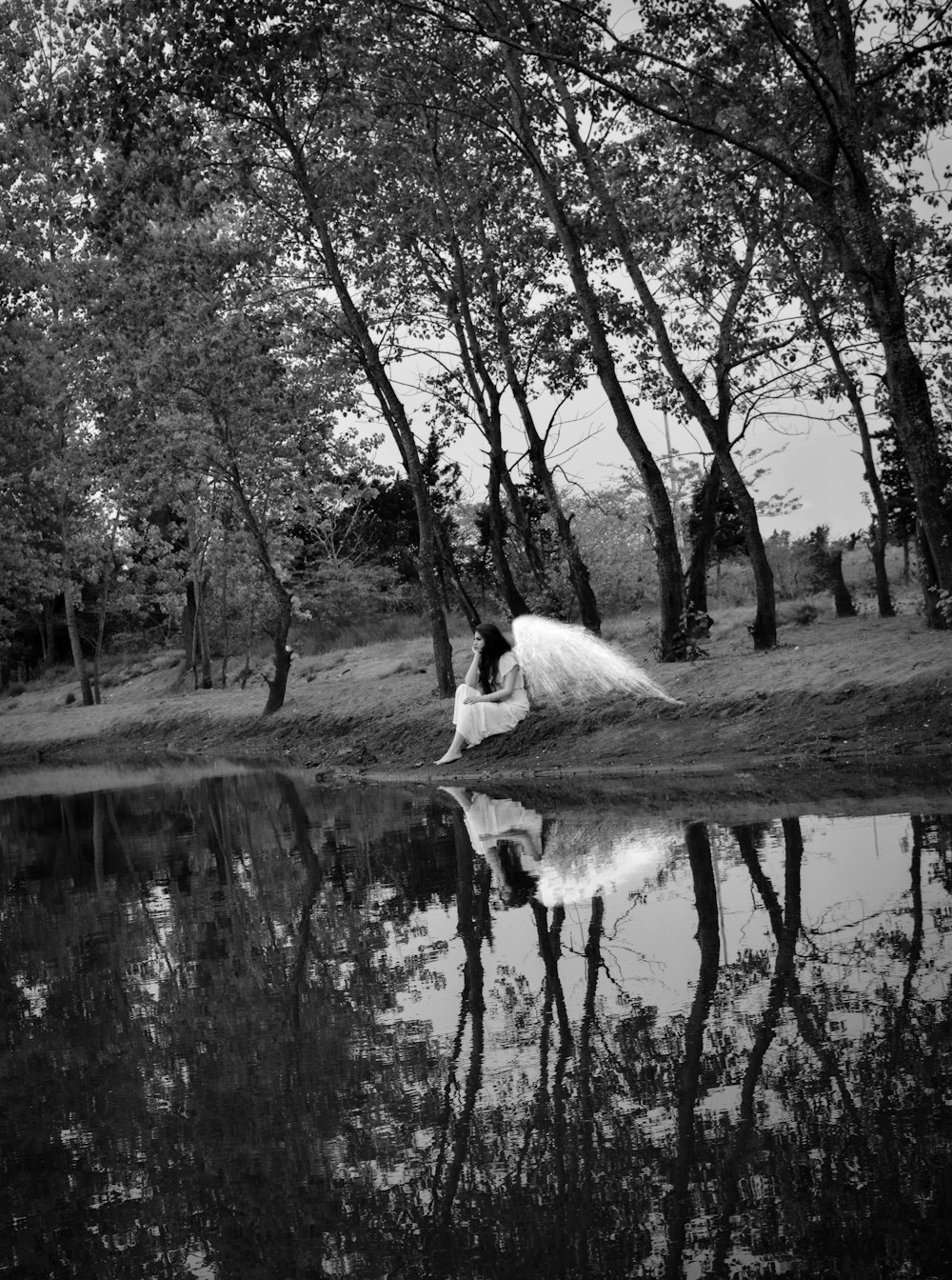 grayscale photography of female angel sitting beside body of water