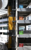 An industrial elevator system in a multi-story parking garage with clearly marked floors in bold colors: blue for the fifth floor, orange for the fourth, green for the third, and red for the second. The structure features concrete beams and glass panels allowing natural light. Yellow elevator cars add a pop of color to the otherwise grey and silver setting, and a few people can be seen on the floors.