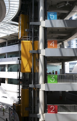 An industrial elevator system in a multi-story parking garage with clearly marked floors in bold colors: blue for the fifth floor, orange for the fourth, green for the third, and red for the second. The structure features concrete beams and glass panels allowing natural light. Yellow elevator cars add a pop of color to the otherwise grey and silver setting, and a few people can be seen on the floors.