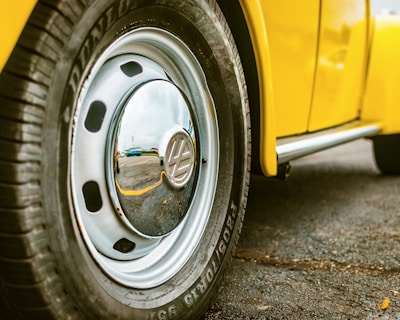 A close-up view of a car wheel featuring a shiny chrome hubcap and a tire with visible tread pattern. The vehicle appears to be yellow, and the surface beneath is asphalt. The reflection in the hubcap suggests an outdoor setting.