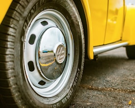 A close-up view of a car wheel featuring a shiny chrome hubcap and a tire with visible tread pattern. The vehicle appears to be yellow, and the surface beneath is asphalt. The reflection in the hubcap suggests an outdoor setting.