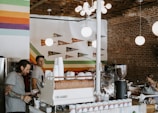 A cozy coffee shop interior with exposed brick walls and colorful stripes painted on a section of the wall. Several round, white pendant lights hang from the ceiling. Two men are behind the counter, one of them holding a cup. The counter has a coffee machine, cups, and various supplies. Wall decorations include small pennants hanging above the seating area on the left. One woman is sitting at a table, working on a laptop.