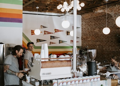 A cozy coffee shop interior with exposed brick walls and colorful stripes painted on a section of the wall. Several round, white pendant lights hang from the ceiling. Two men are behind the counter, one of them holding a cup. The counter has a coffee machine, cups, and various supplies. Wall decorations include small pennants hanging above the seating area on the left. One woman is sitting at a table, working on a laptop.