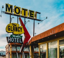 A vintage motel sign with distinct retro typography and colors stands in front of an old motel building. The sign includes bold, weathered letters spelling 'Motel' and 'The Glancy Motor Hotel,' adorned with large arrows and mounted on metal posts. The motel building shows signs of age with peeling paint and a nostalgic architectural style. The sky is partly cloudy, adding to the image's timeless feel.