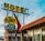 A vintage motel sign with distinct retro typography and colors stands in front of an old motel building. The sign includes bold, weathered letters spelling 'Motel' and 'The Glancy Motor Hotel,' adorned with large arrows and mounted on metal posts. The motel building shows signs of age with peeling paint and a nostalgic architectural style. The sky is partly cloudy, adding to the image's timeless feel.