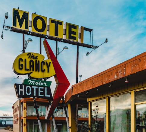 A vintage motel sign with distinct retro typography and colors stands in front of an old motel building. The sign includes bold, weathered letters spelling 'Motel' and 'The Glancy Motor Hotel,' adorned with large arrows and mounted on metal posts. The motel building shows signs of age with peeling paint and a nostalgic architectural style. The sky is partly cloudy, adding to the image's timeless feel.
