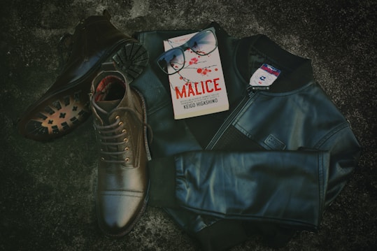 A pair of dark leather boots is laying on a textured surface alongside a sleek black leather jacket. An open book with a red and black cover titled 'Malice' is placed next to black sunglasses on the jacket. The scene conveys a sense of mystery and style.