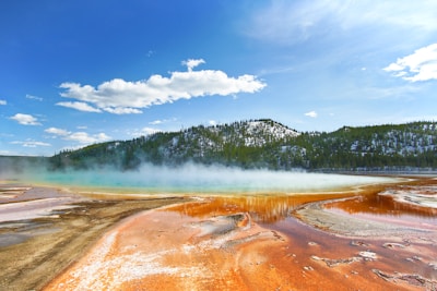 A vibrant hot spring with a wide spectrum of colors including orange, yellow, and light blue, surrounded by a forested hill under a bright blue sky with scattered clouds. The water emits steam, creating a mystical effect.