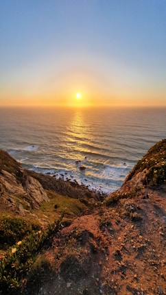 A breathtaking view of a sunset over the ocean, with the sun casting golden reflections on the water. In the foreground, there is a rocky cliff with some vegetation, leading down to a beach with waves crashing against the shore.