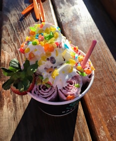 A serving of rolled ice cream topped with whipped cream and colorful, crunchy cereal pieces. A whole strawberry is placed on the side, and a pink wafer stick is also inserted into the ice cream. The dessert is in a cup on a wooden surface with sunlight illuminating the dish.