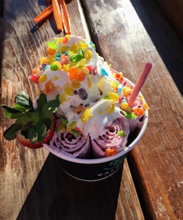 A serving of rolled ice cream topped with whipped cream and colorful, crunchy cereal pieces. A whole strawberry is placed on the side, and a pink wafer stick is also inserted into the ice cream. The dessert is in a cup on a wooden surface with sunlight illuminating the dish.