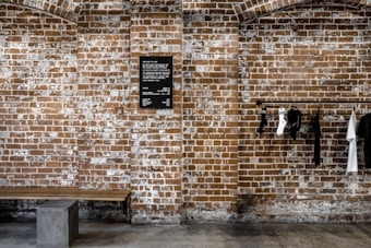 An exposed brick wall features a black menu board mounted on its surface. To the right, a clothing rack displays a selection of black and white garments, including shirts and socks. Below the board, a minimalist bench with a wooden top and a concrete base is positioned against the wall.