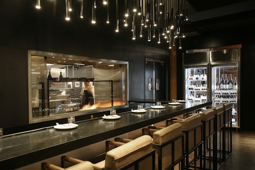 A modern kitchen with a chef working at a grill, visible through a large glass window. The kitchen is equipped with professional appliances and has a warm, inviting glow. There is a sleek countertop with cushioned chairs set for dining, and overhead lighting features numerous small, pendant lights. On one side, a glass refrigerator displays a variety of wine bottles.
