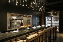 A modern kitchen with a chef working at a grill, visible through a large glass window. The kitchen is equipped with professional appliances and has a warm, inviting glow. There is a sleek countertop with cushioned chairs set for dining, and overhead lighting features numerous small, pendant lights. On one side, a glass refrigerator displays a variety of wine bottles.