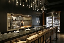 A modern kitchen with a chef working at a grill, visible through a large glass window. The kitchen is equipped with professional appliances and has a warm, inviting glow. There is a sleek countertop with cushioned chairs set for dining, and overhead lighting features numerous small, pendant lights. On one side, a glass refrigerator displays a variety of wine bottles.